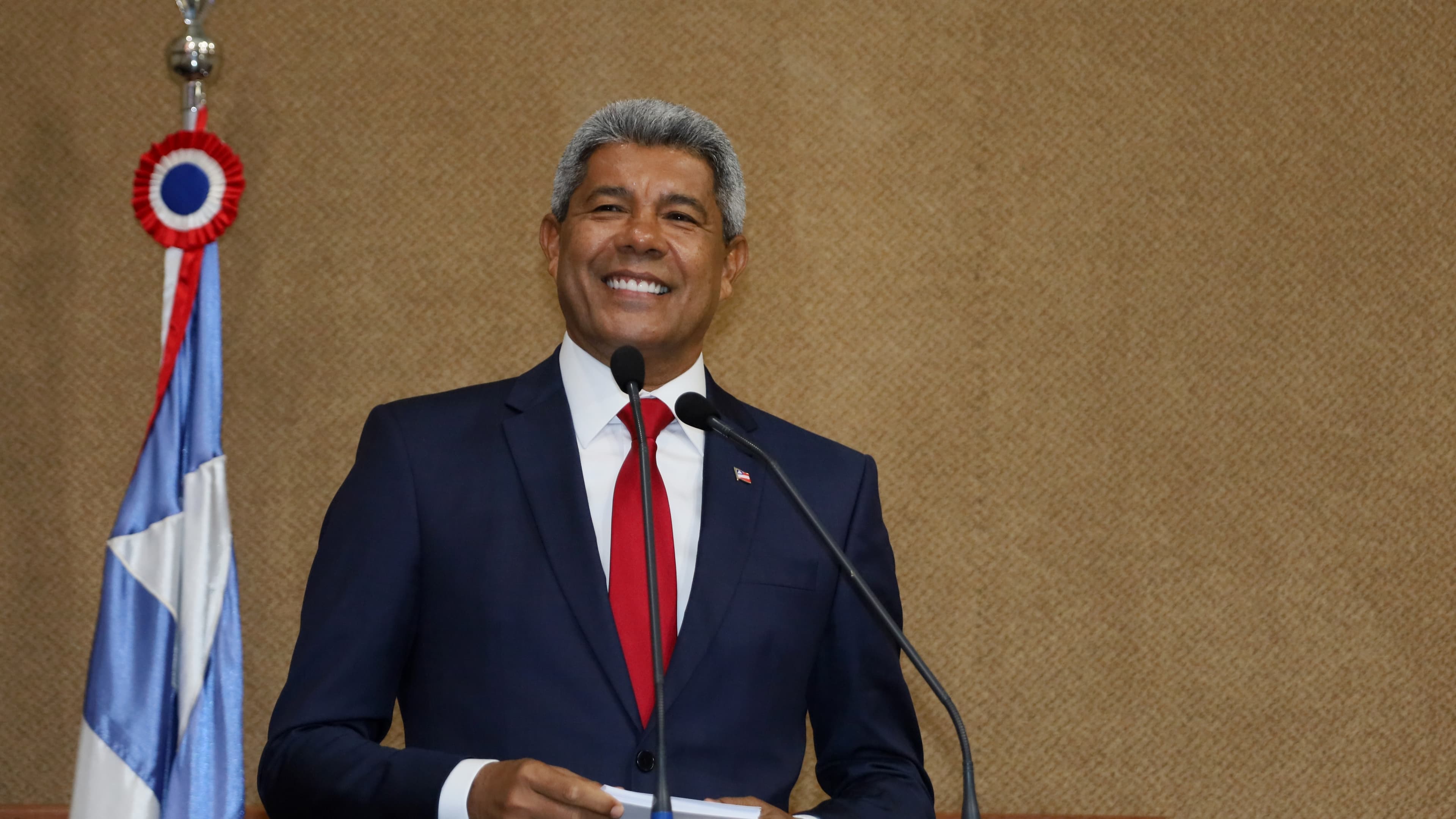 Jerônimo Rodrigues, de terno azul e gravata vermelha, sorri ao microfone ao lado da bandeira da Bahia, em foto de arquivo