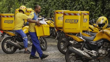 Carteiros dos Correios com uniforme amarelo carregam caixas ao lado de motos Sedex estacionadas