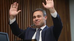 André Mendonça sorri com as mãos levantadas em evento oficial, de terno azul-marinho