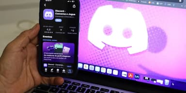 Homem segura um celular com o aplicativo do Discord aberto, com notebook ao fundo mostrando o logotipo do Discord
