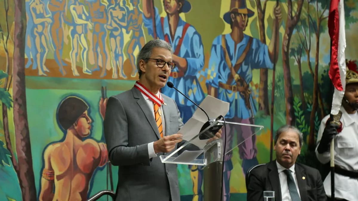 Romeu Zema discursa em cerimônia usando medalha no peito, ao lado de outra autoridade sentada