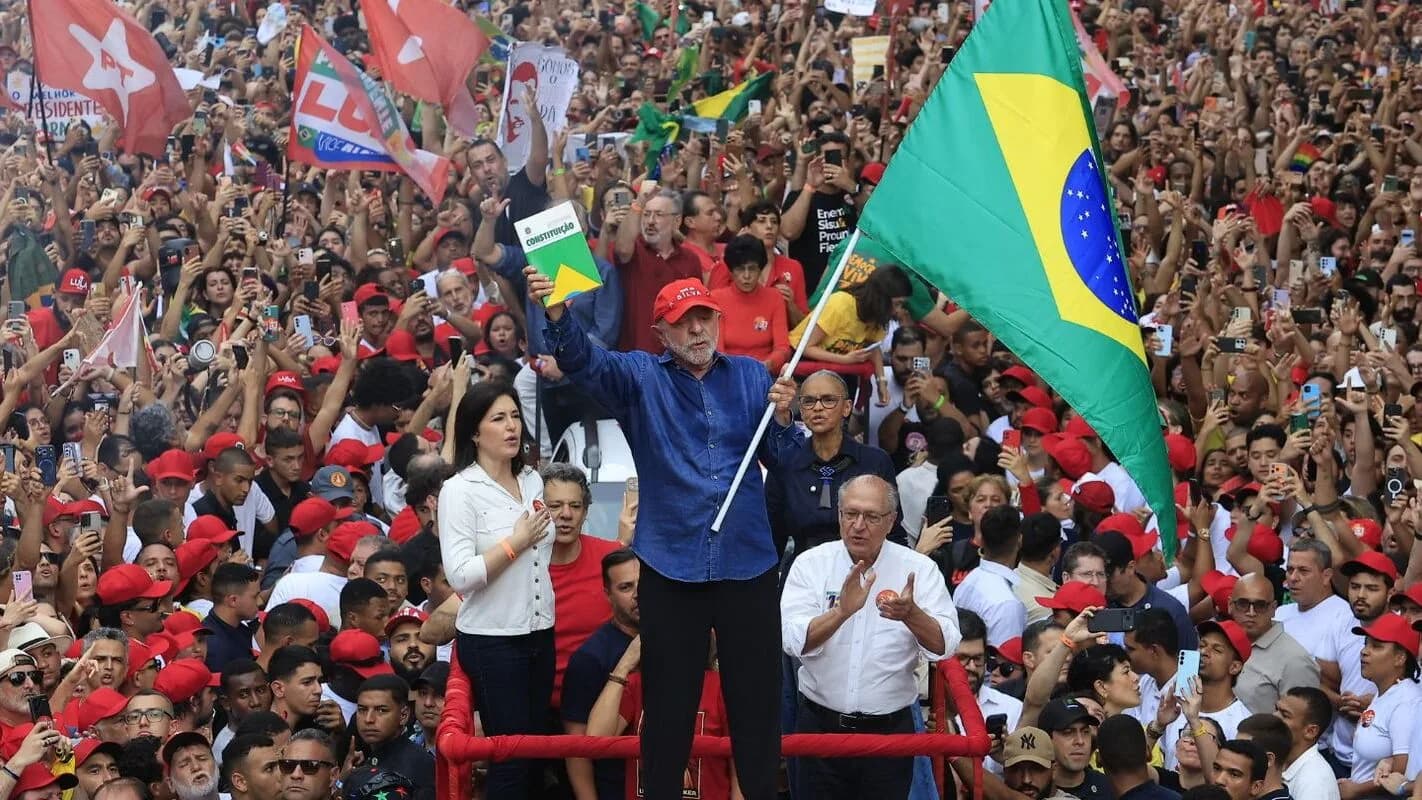 Lula ao lado de Alckmin ergue a bandeira do Brasil e um exemplar da Constituição em ato com multidão