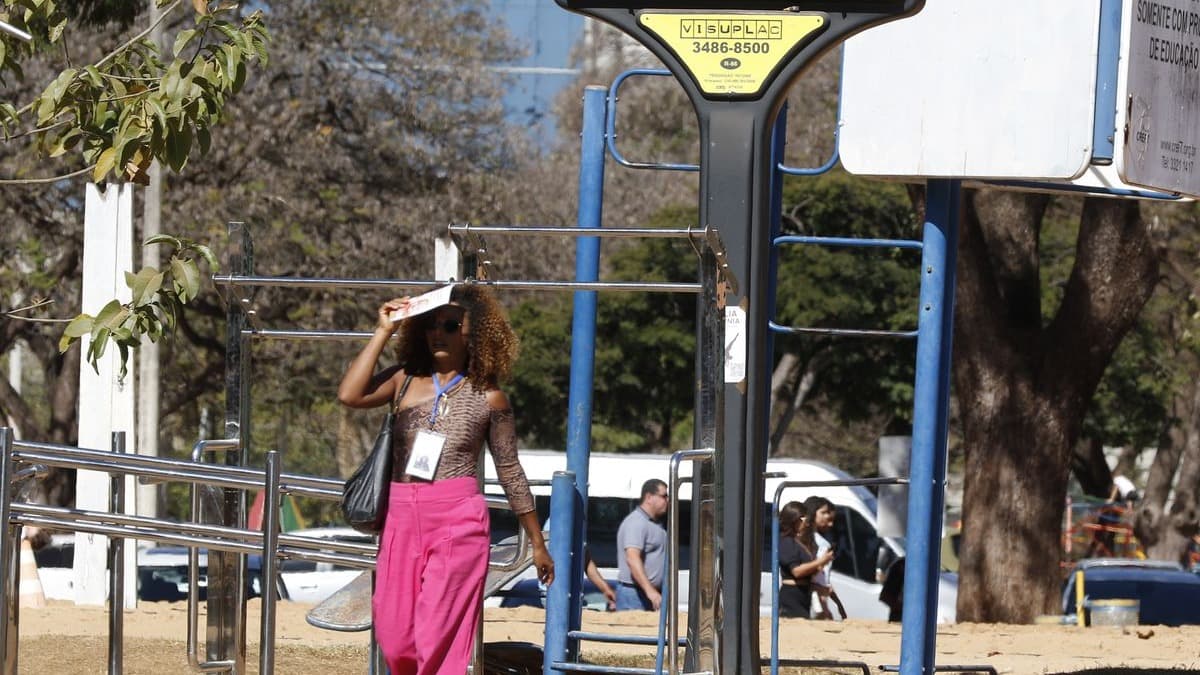 Mulher se protege do sol com uma pasta sobre a cabeça ao caminhar por área de lazer com solo seco e árvores sem folhas, em dia de tempo seco; imagem de arquivo