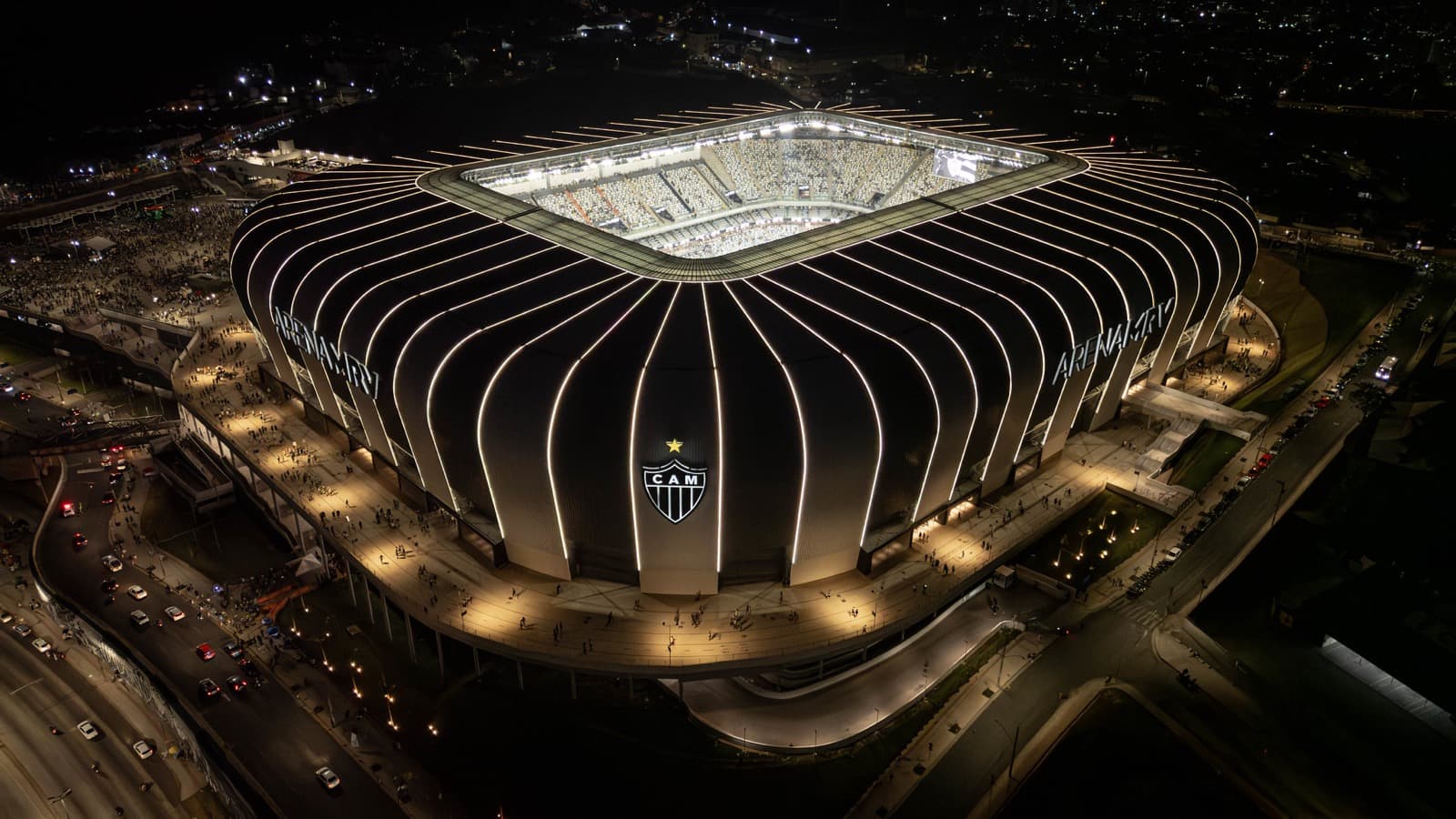 Imagem aérea de arquivo da Arena MRV, estádio do Atlético-MG em Belo Horizonte, iluminada à noite, com o escudo do clube na fachada e torcedores na esplanada