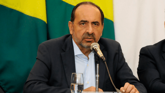 Alexandre Kalil discursa em coletiva, vestindo terno escuro, diante de microfone e bandeiras