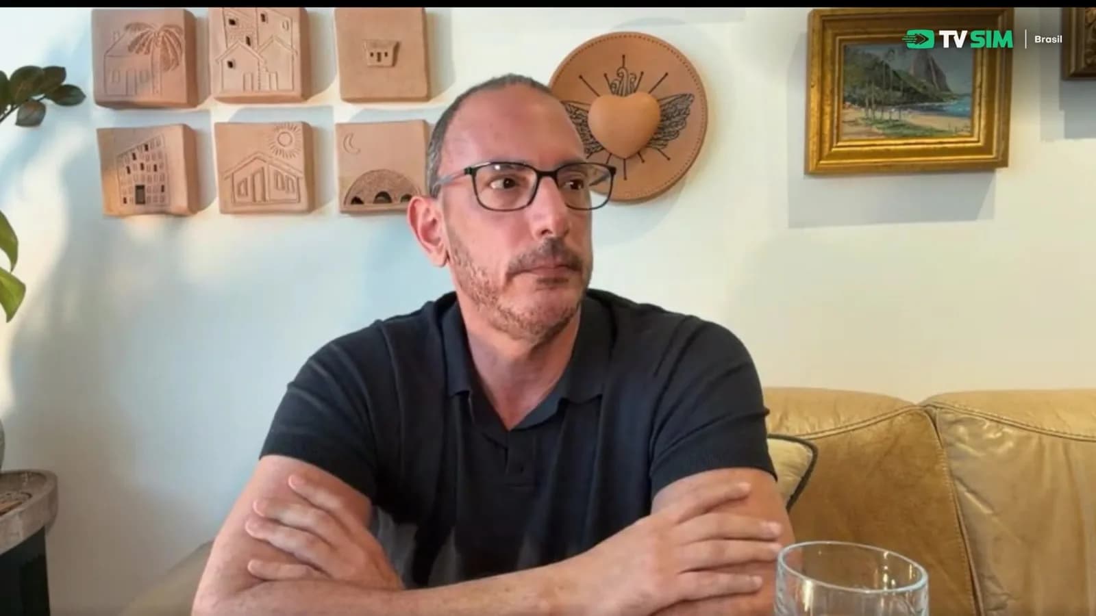 O consultor político Abel Lumer, de óculos e camisa polo escura, com os braços cruzados sobre a mesa durante entrevista em um sofá de couro, com a parede decorada ao fundo