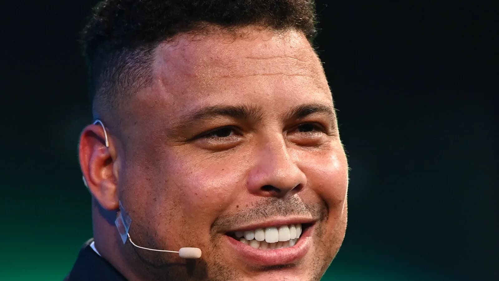 Ronaldo Nazário sorri durante evento, usando microfone de lapela e terno escuro