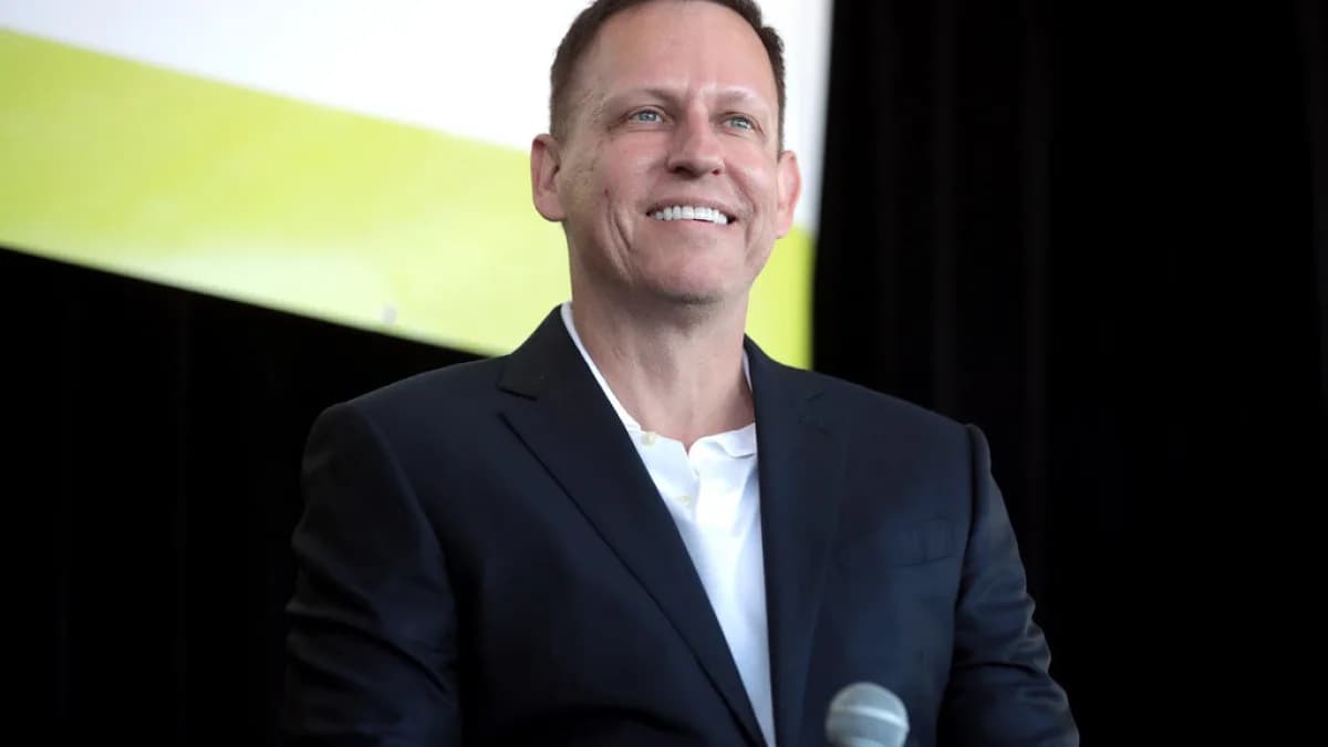 Peter Thiel sorri durante evento, de terno escuro, ao lado de um microfone