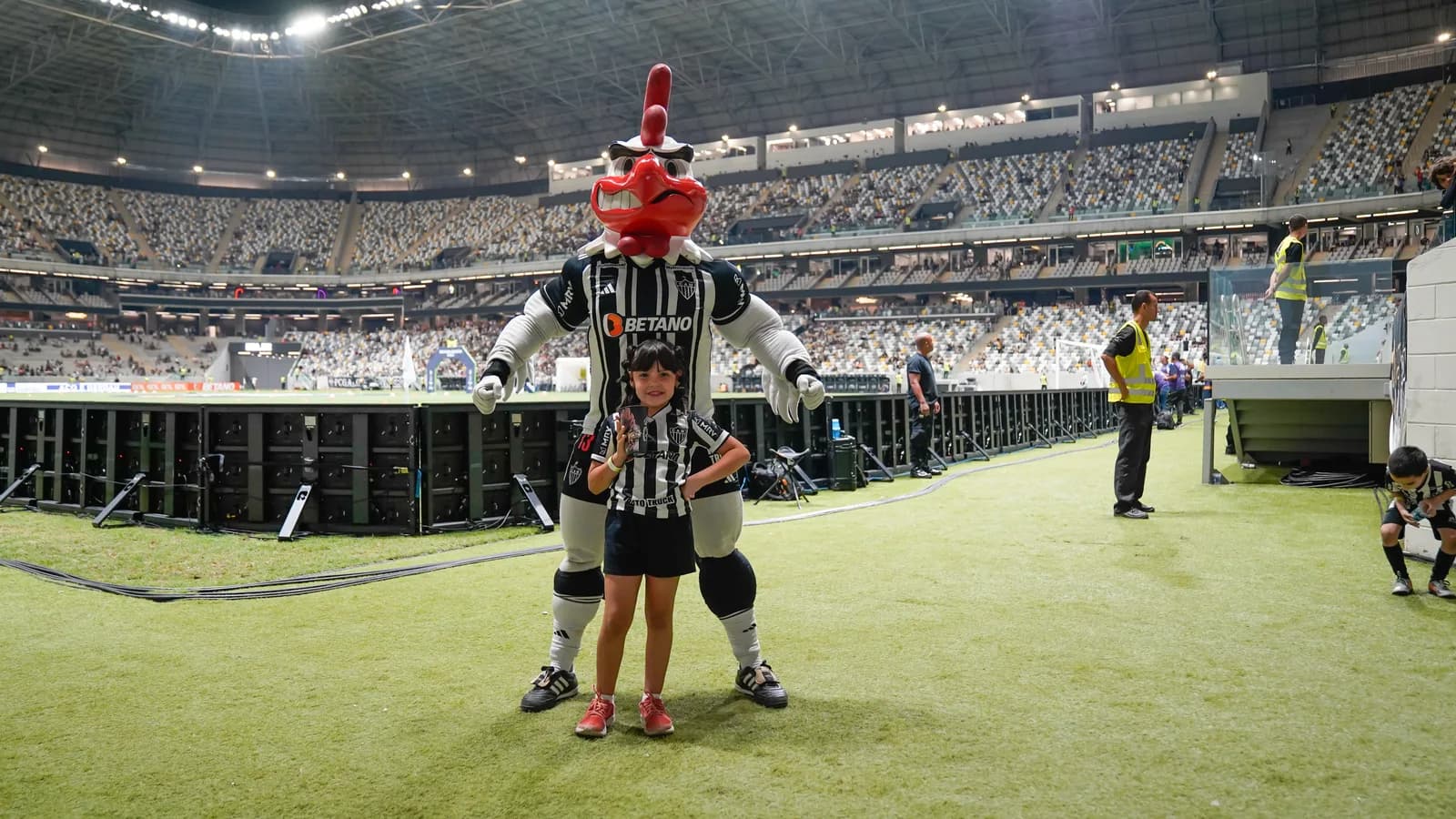 Mascote do Atlético-MG, o Galo Doido, ao lado de uma criança no gramado da Arena MRV lotada