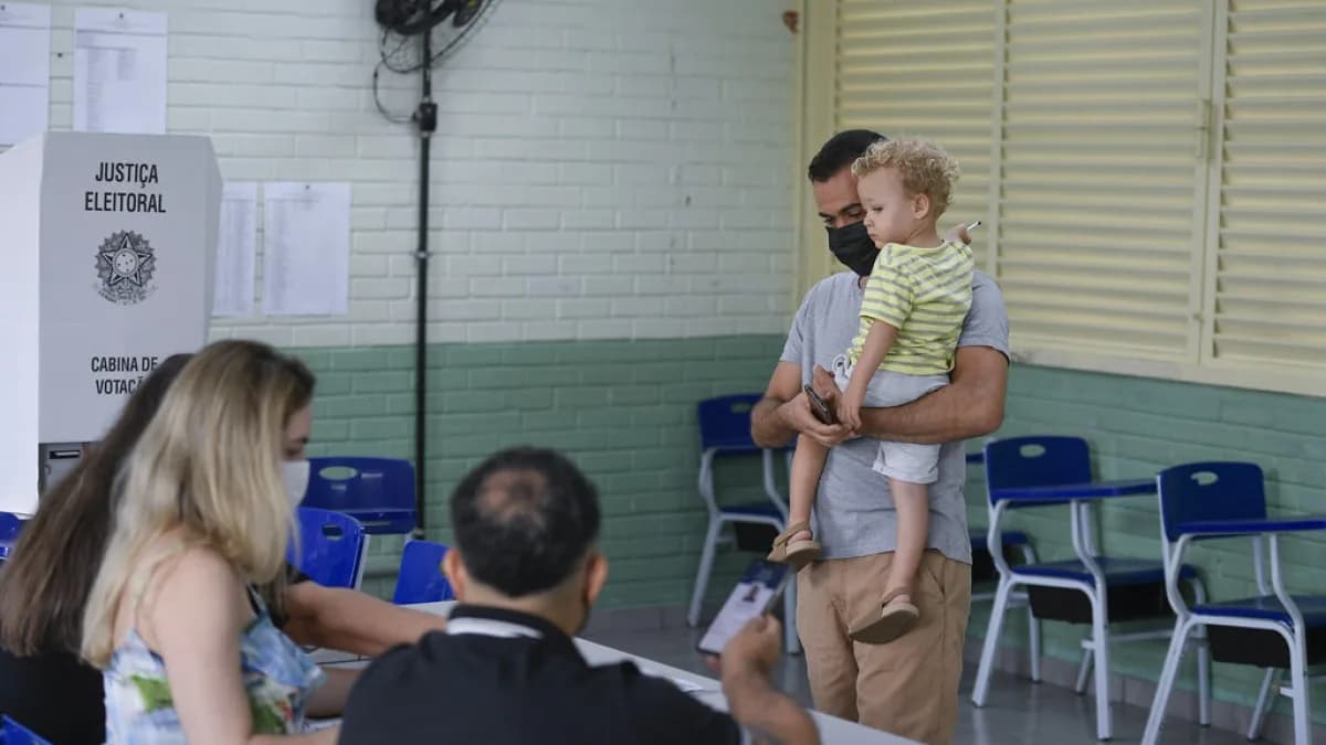 Eleitor com criança no colo em cabine de votação, acompanhado por mesário em seção eleitoral