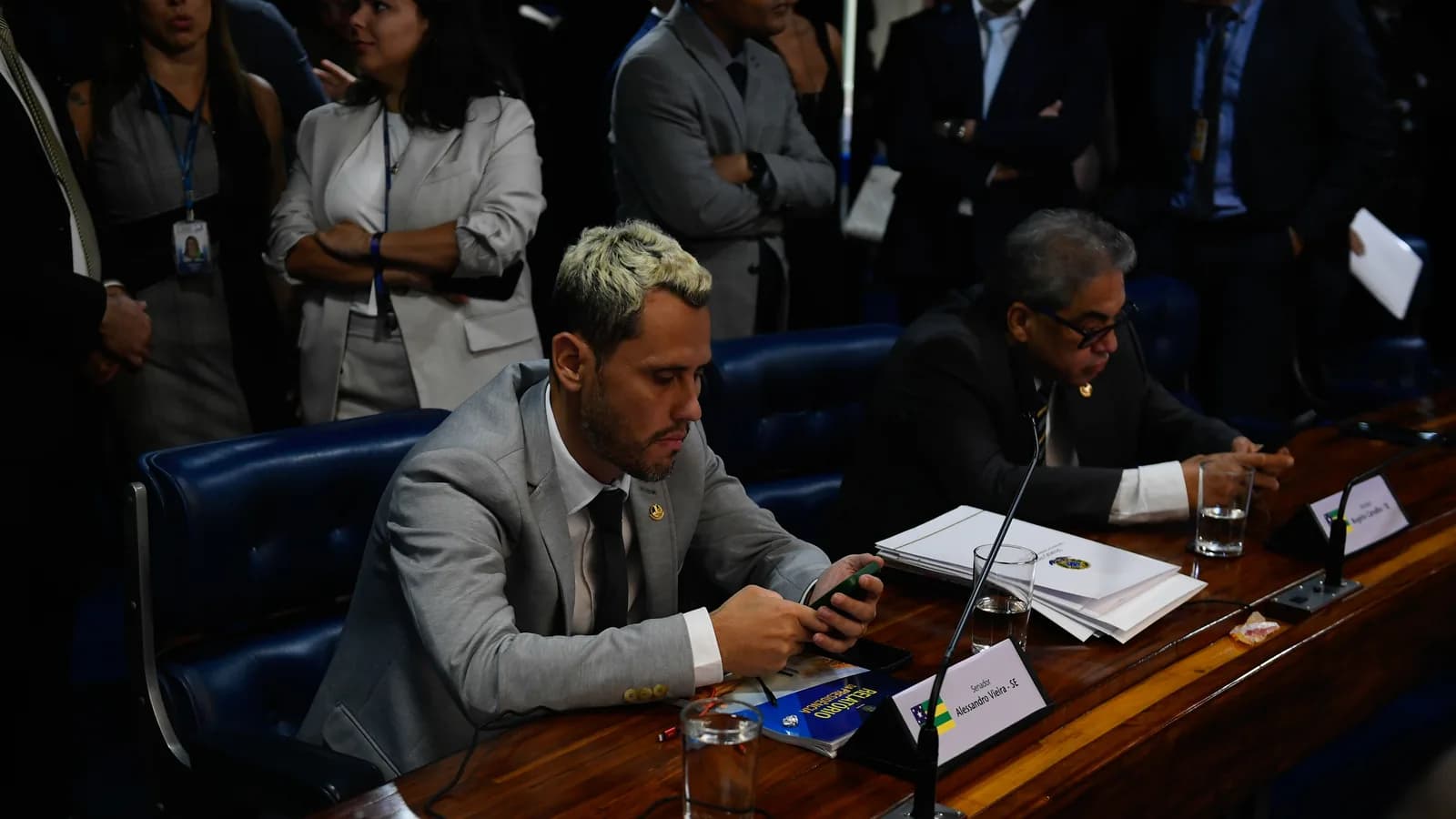 Cleitinho Azevedo sentado em mesa de comissão no Senado, olhando para o celular