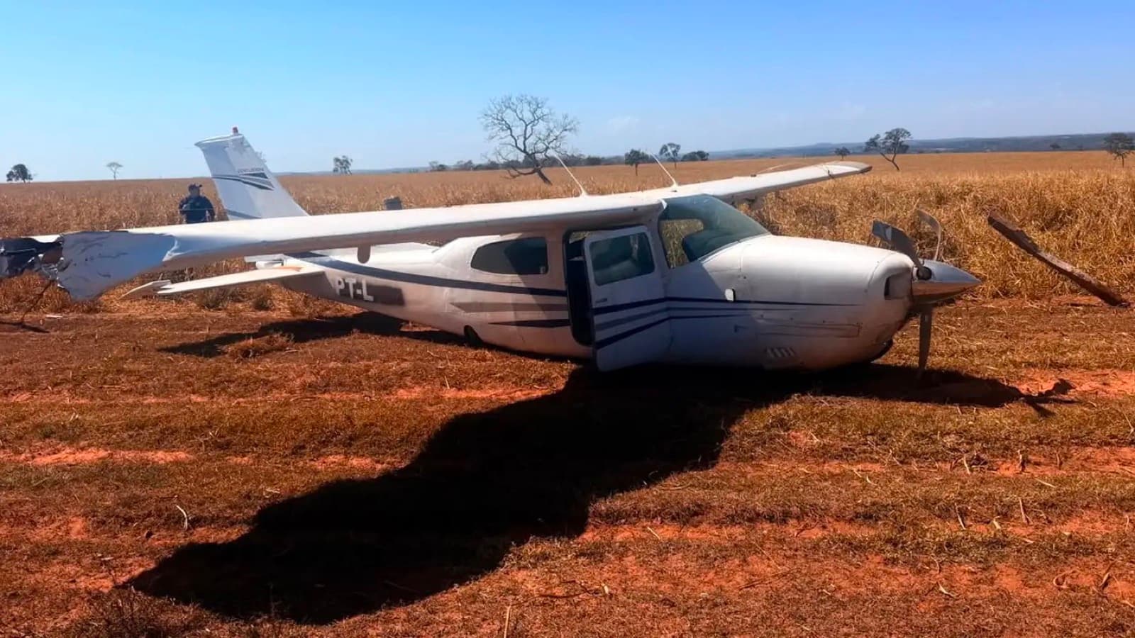 Avião monomotor branco pousado à força em lavoura de área rural, com a hélice arrancada e a porta aberta; ao fundo, dois agentes e o horizonte de plantação