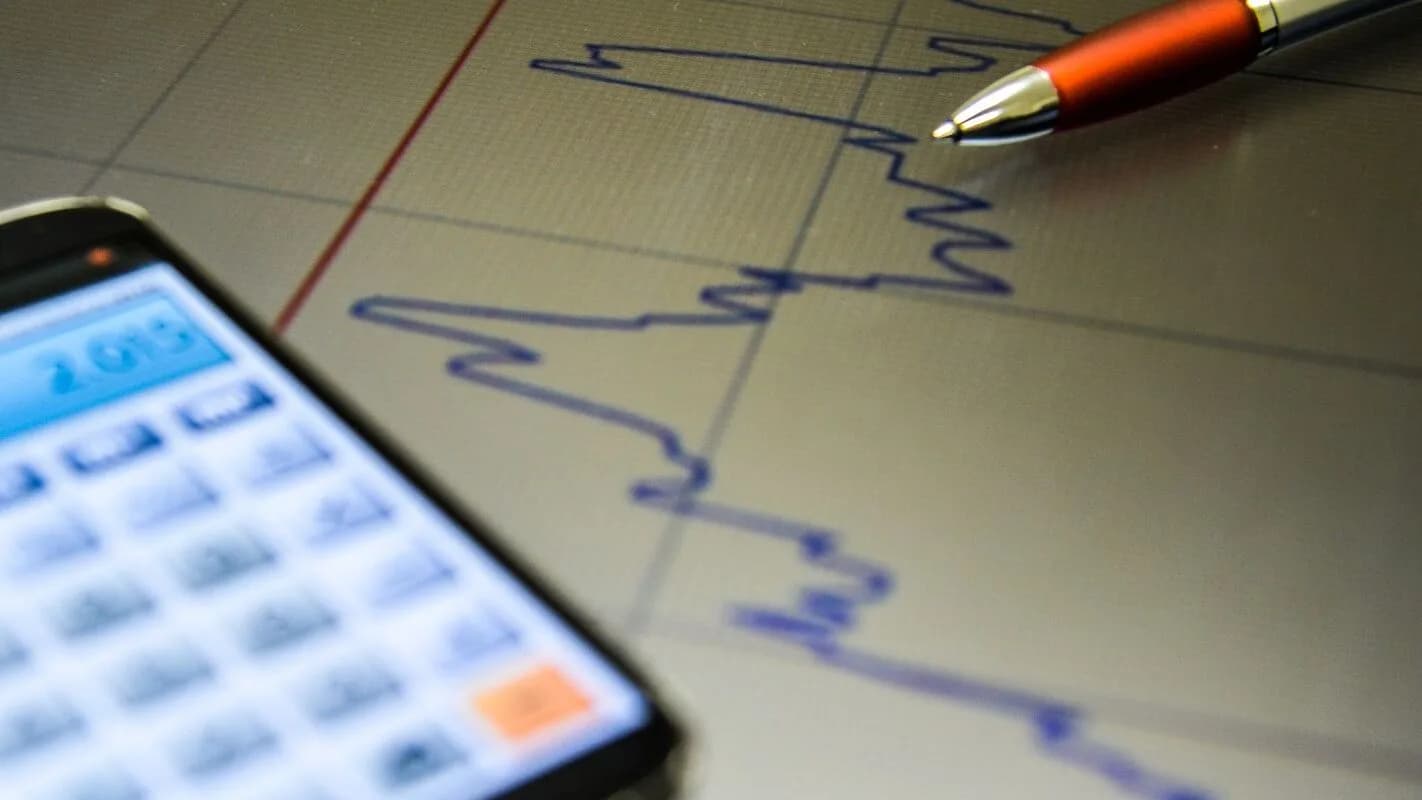 Gráfico de mercado financeiro em tela de celular ao lado de caneta sobre relatório impresso