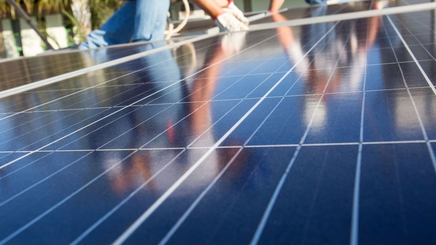 Técnico com luvas instala painéis solares fotovoltaicos, vistos de perto e refletindo o céu