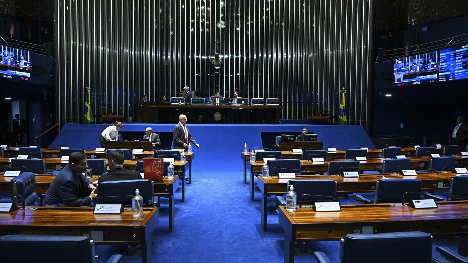 Vista do plenário do Senado Federal, com bancadas de madeira e cadeiras azuis em primeiro plano e senadores ao fundo durante sessão