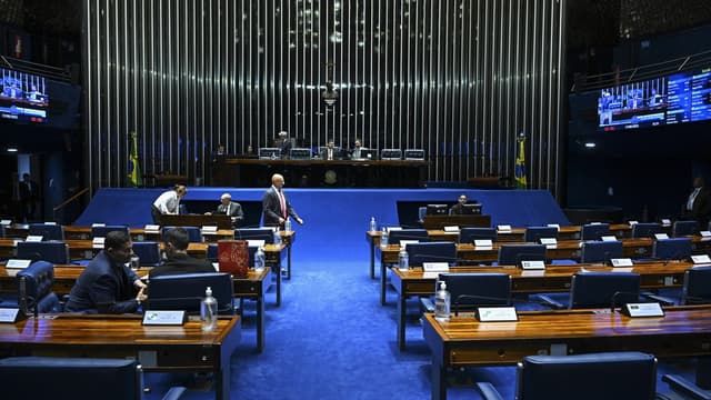 Vista do plenário do Senado Federal, com bancadas de madeira e cadeiras azuis em primeiro plano e senadores ao fundo durante sessão