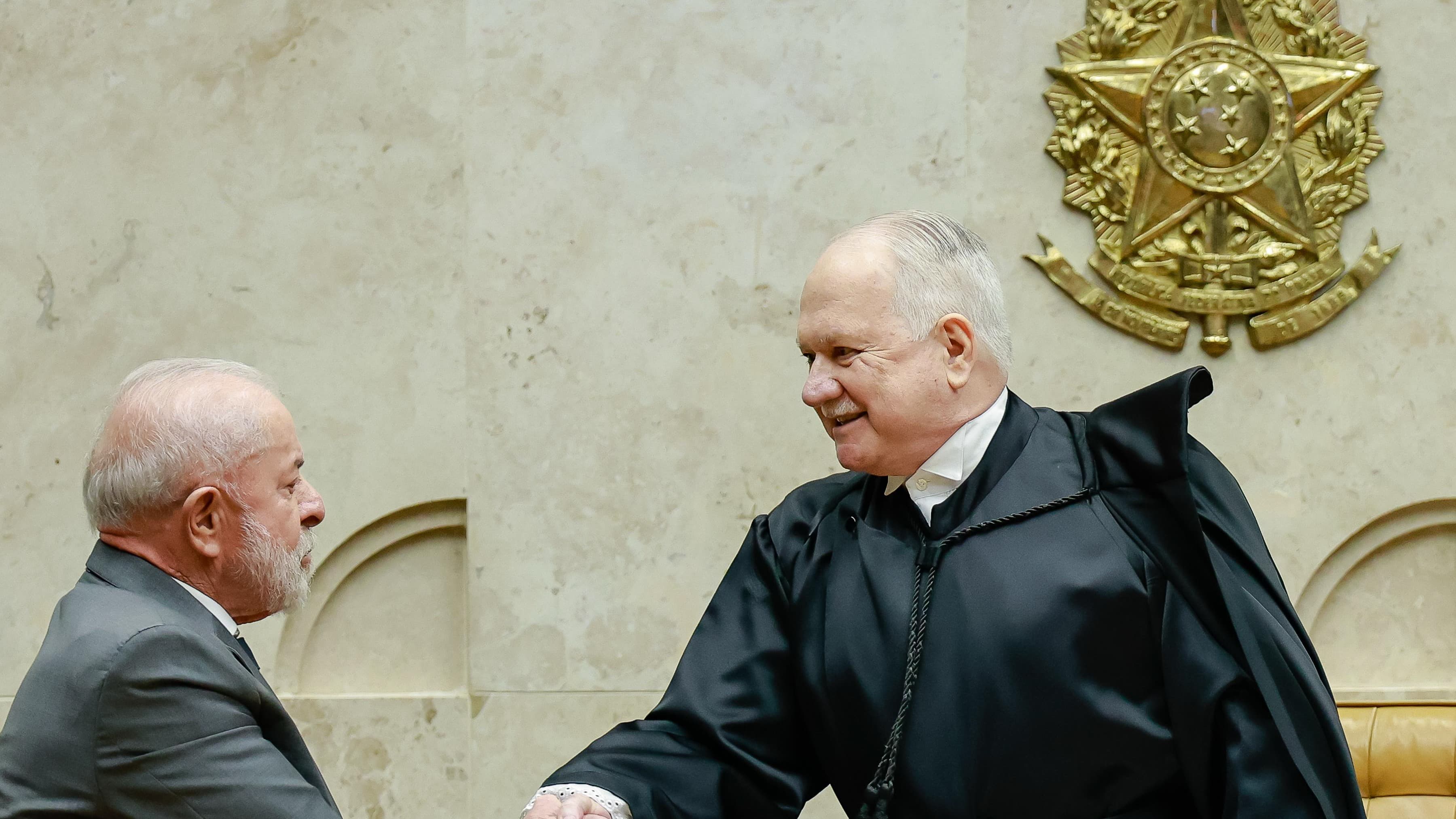 O ministro Edson Fachin, presidente do STF, cumprimenta o presidente Lula no plenário do Supremo, com o brasão da República ao fundo