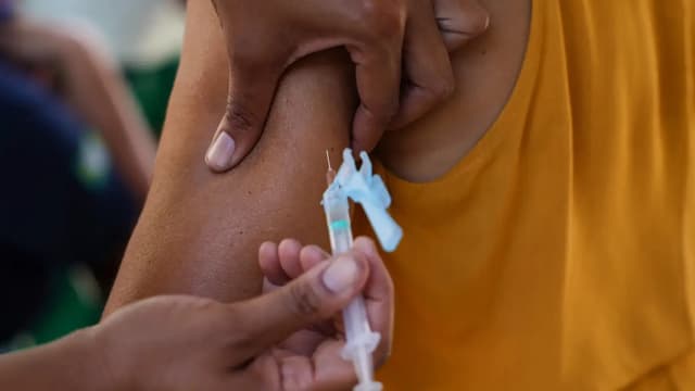 Imagem de arquivo: profissional de saúde aplica vacina no braço de uma pessoa de camiseta amarela, em close, sem rostos identificáveis