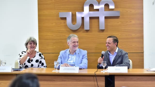Três pessoas sentadas a uma mesa de coletiva de imprensa, sorrindo, diante de um painel de madeira com o logotipo da UFF; o homem à direita fala ao microfone