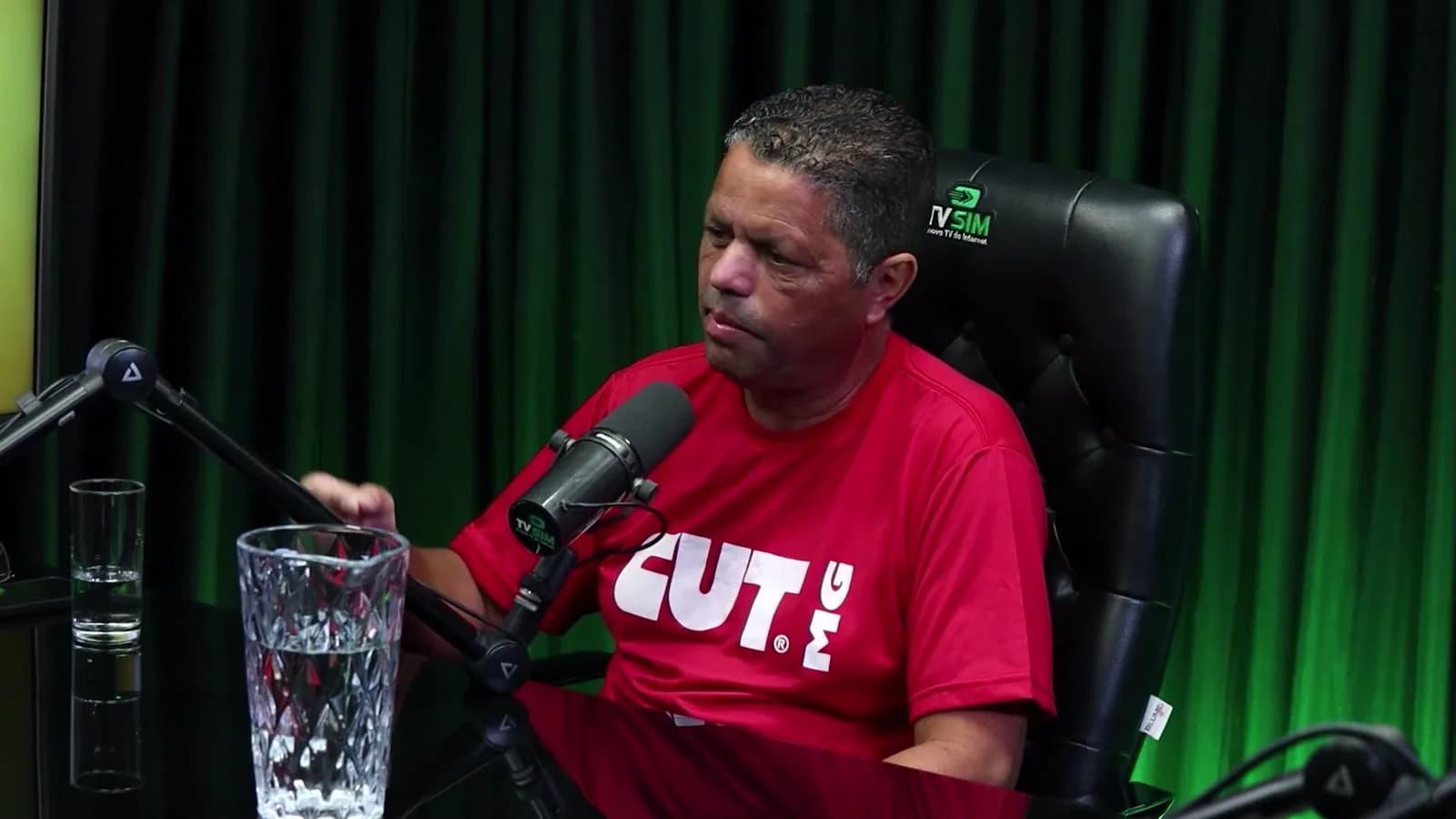 Jairon Nogueira Filho, presidente da CUT em Minas Gerais, de camiseta vermelha da CUT-MG, fala gesticulando em estúdio com cortina verde ao fundo, ao lado de um copo d'água na mesa.