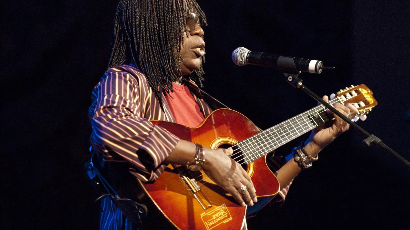 Milton Nascimento canta e toca violão em show, com trancinhas e jaqueta listrada