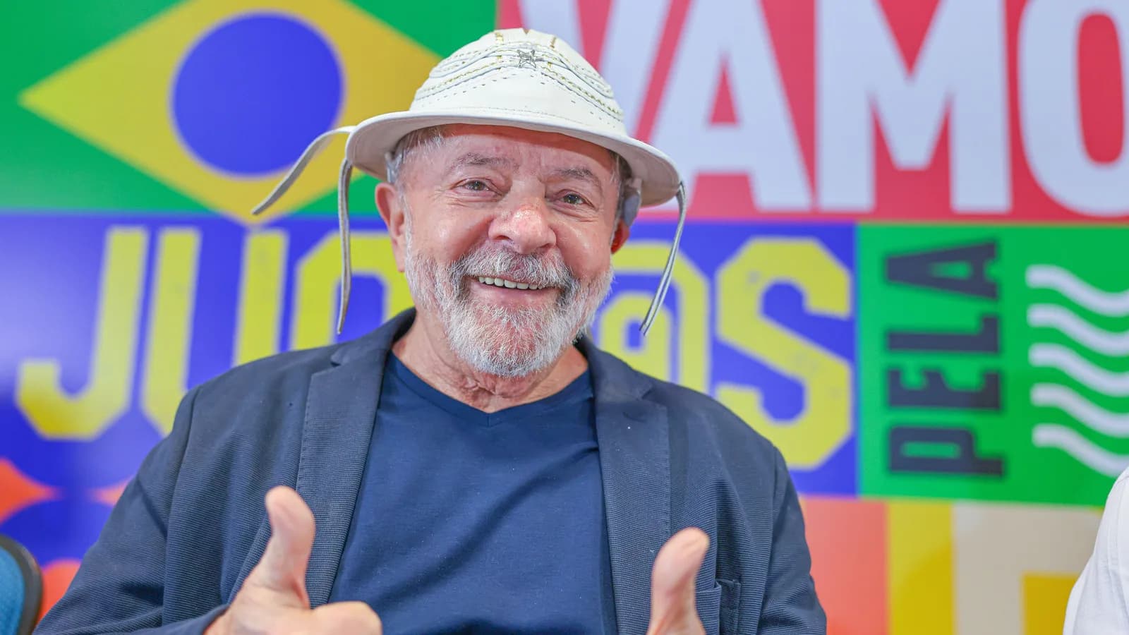 Lula sorri e faz sinal de positivo com as duas mãos em evento de campanha, com painel colorido ao fundo