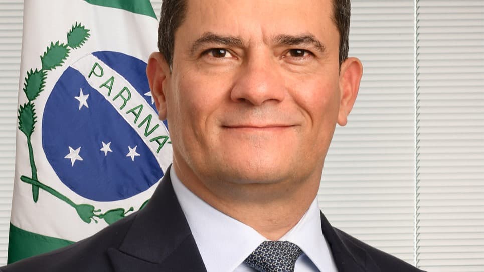 Retrato de Sergio Moro sorrindo, de terno, com a bandeira do Paraná ao fundo