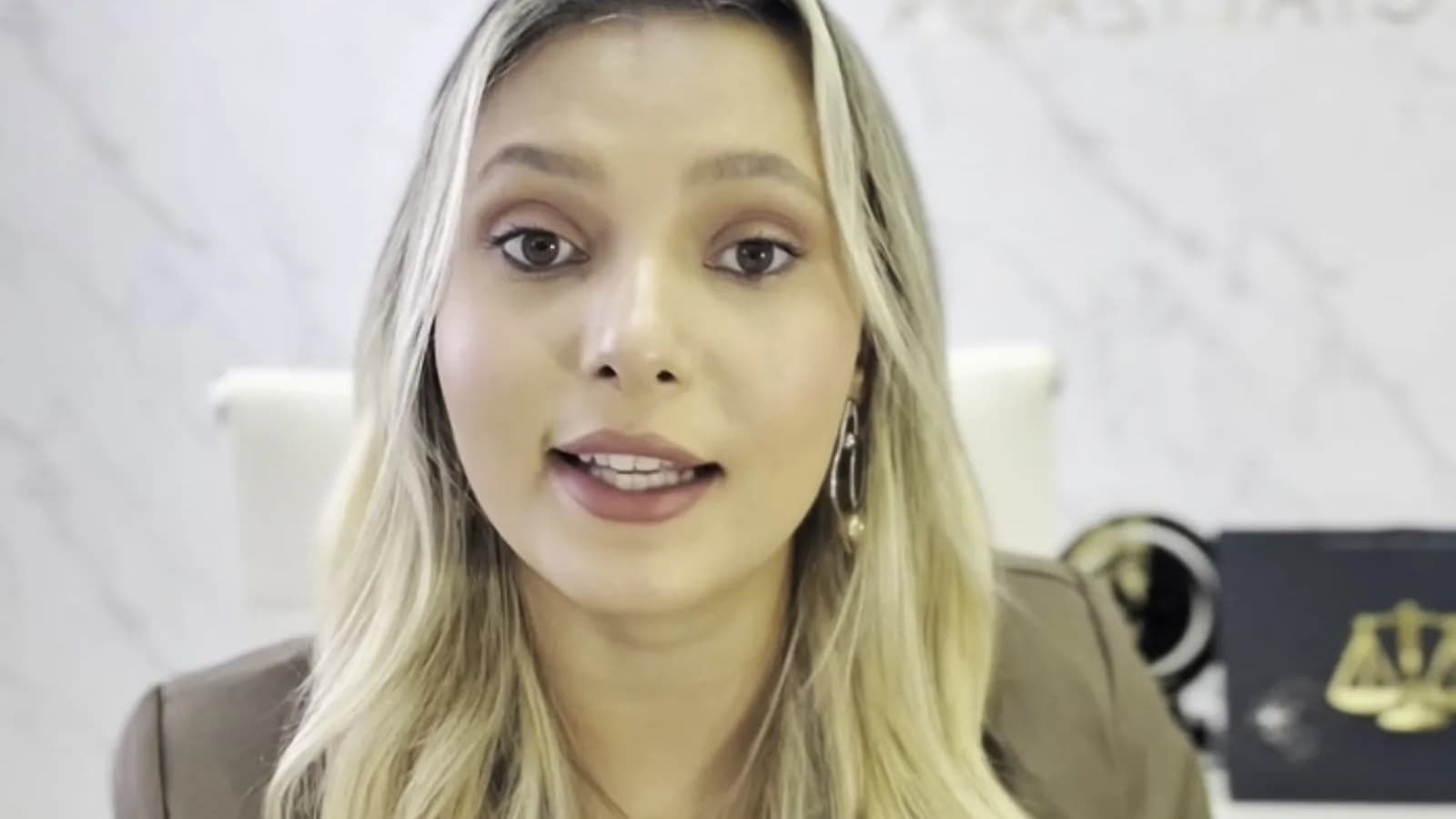 Advogada Isabela Costa fala sobre controle de celular em relacionamentos durante vídeo, com fundo de mármore e símbolo da balança da Justiça