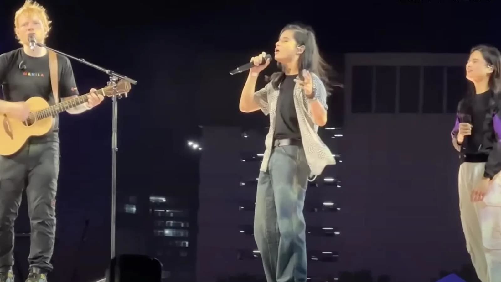 Ed Sheeran toca violão e canta ao lado de outros artistas em show, com prédios iluminados ao fundo