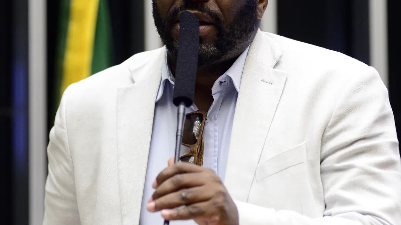 Netinho de Paula discursando ao microfone no plenário da Câmara dos Deputados, vestindo terno branco