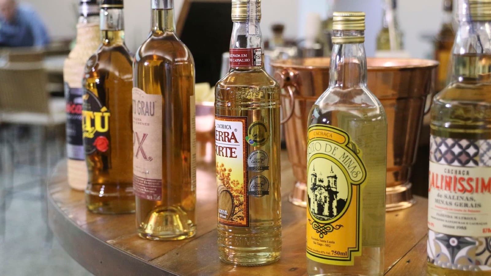 Garrafas de cachaça de marcas mineiras sobre mesa de madeira, com balde de cobre ao fundo; imagem de arquivo