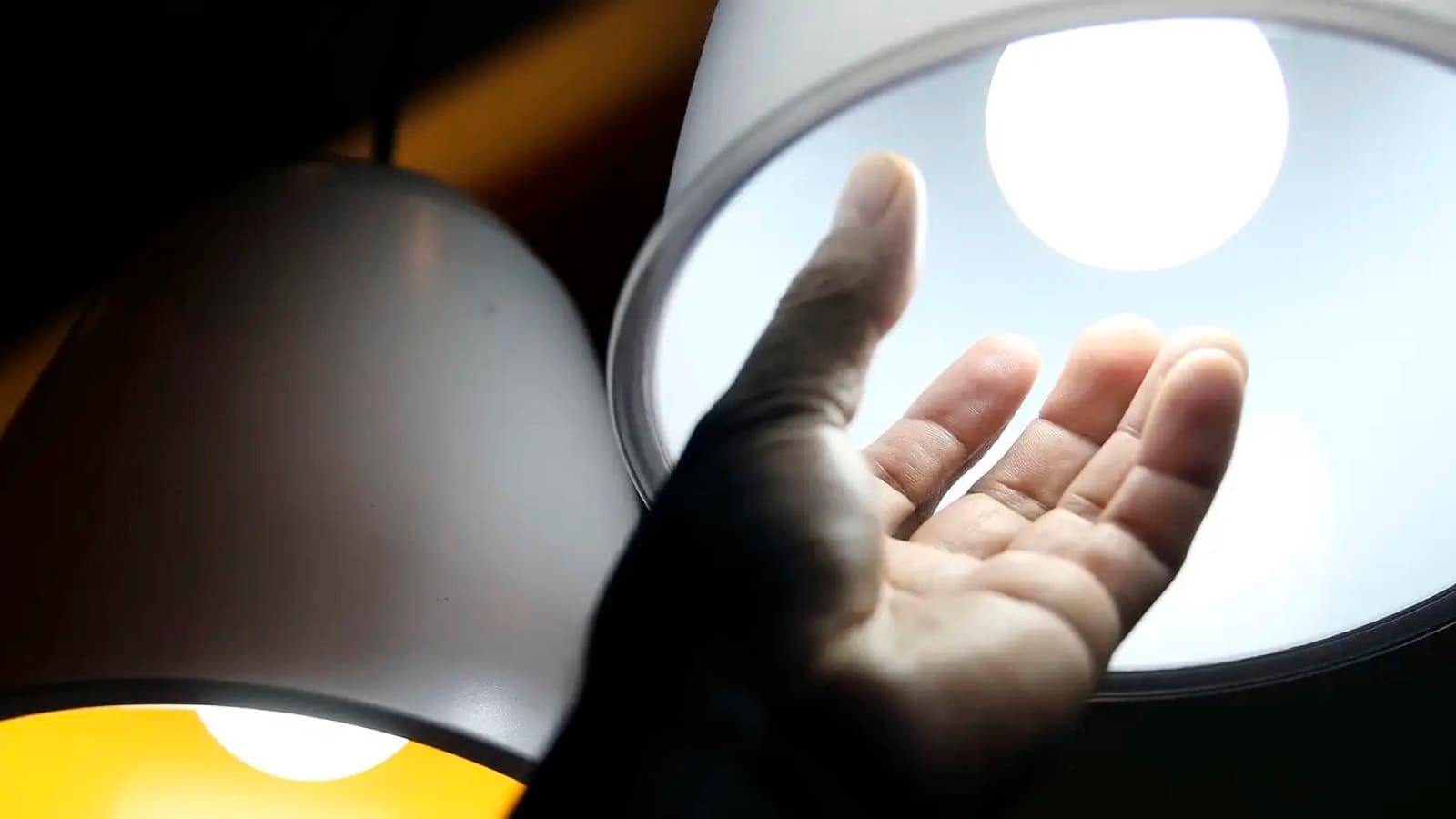 Mão aberta sob luminárias com lâmpadas de LED acesas, em imagem de contexto sobre consumo de energia elétrica