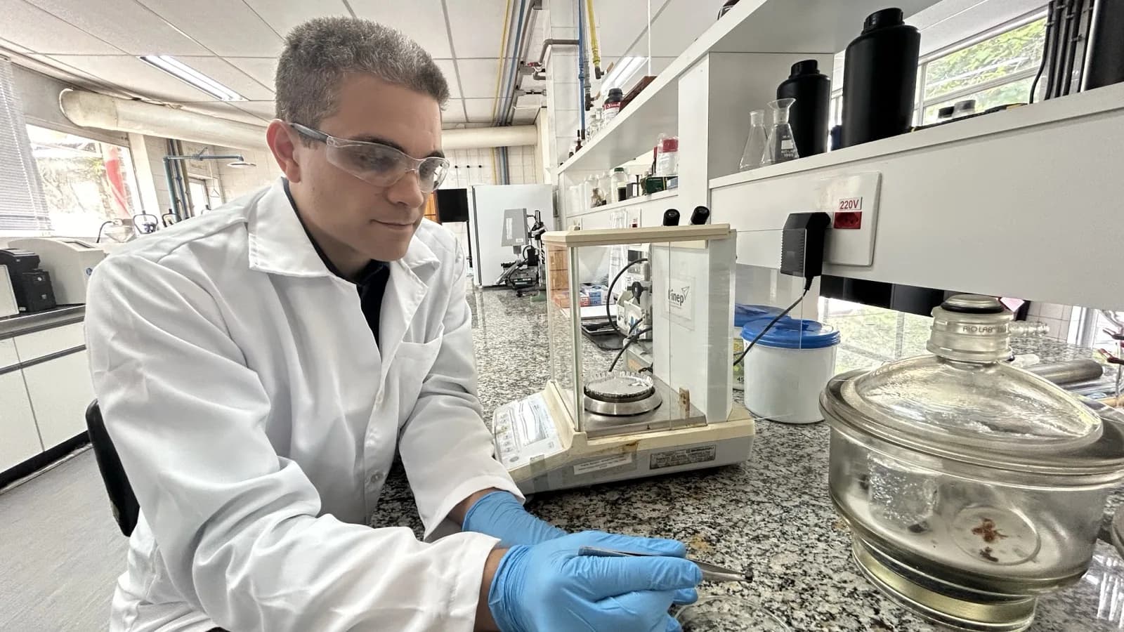 Pesquisador de jaleco branco e luvas manuseia amostra em laboratório com bancada de granito e vidrarias