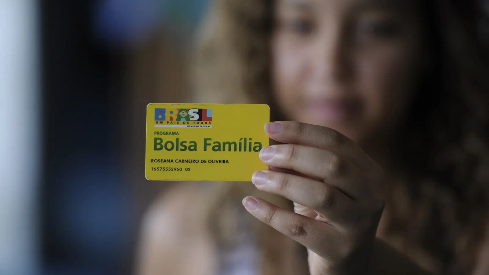 Mulher segura cartão do Programa Bolsa Família em foco, com rosto desfocado ao fundo
