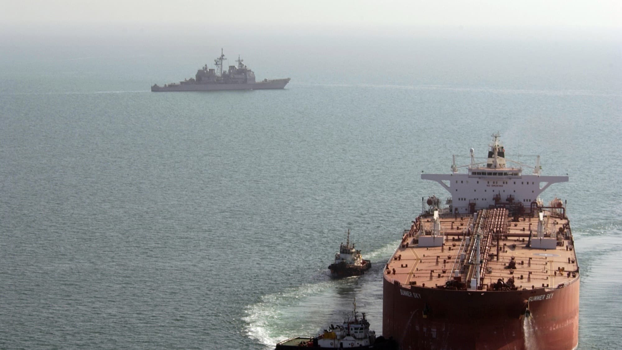 Navio de guerra da Marinha dos EUA patrulha ao fundo enquanto rebocadores guiam um superpetroleiro no Golfo Pérsico