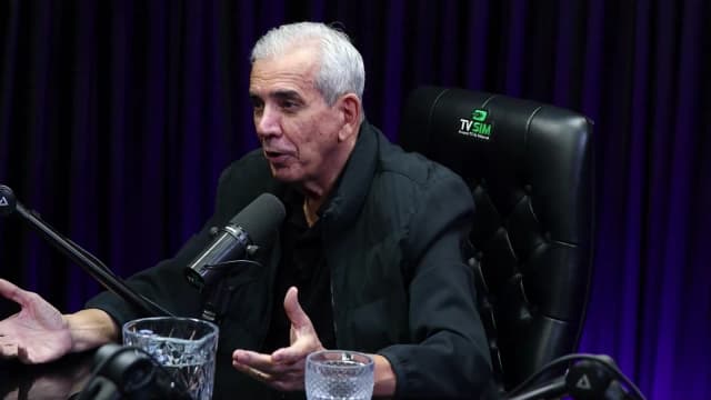 Ricardo Teagano com as duas mãos abertas sobre a mesa do estúdio, gesticulando enquanto explica um ponto da entrevista