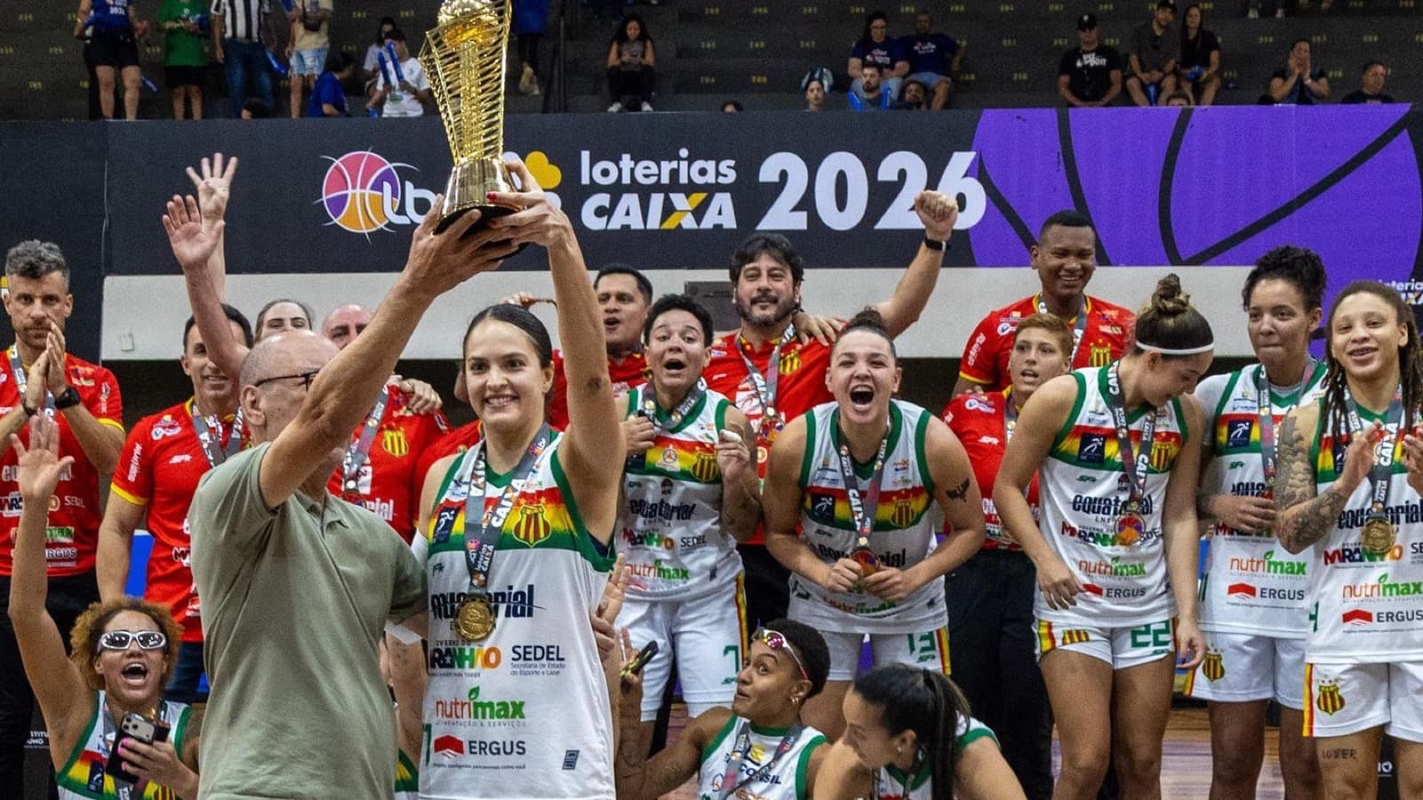 Jogadoras e comissão técnica do Sampaio Basquete comemoram com o troféu erguido, de medalha no peito, diante do painel da Liga de Basquete Feminino 2026 na quadra
