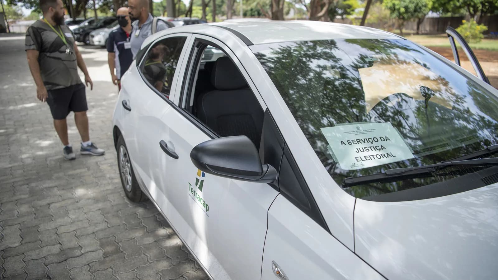 Carro identificado como 'a serviço da Justiça Eleitoral' estacionado em rua arborizada, com homens ao fundo