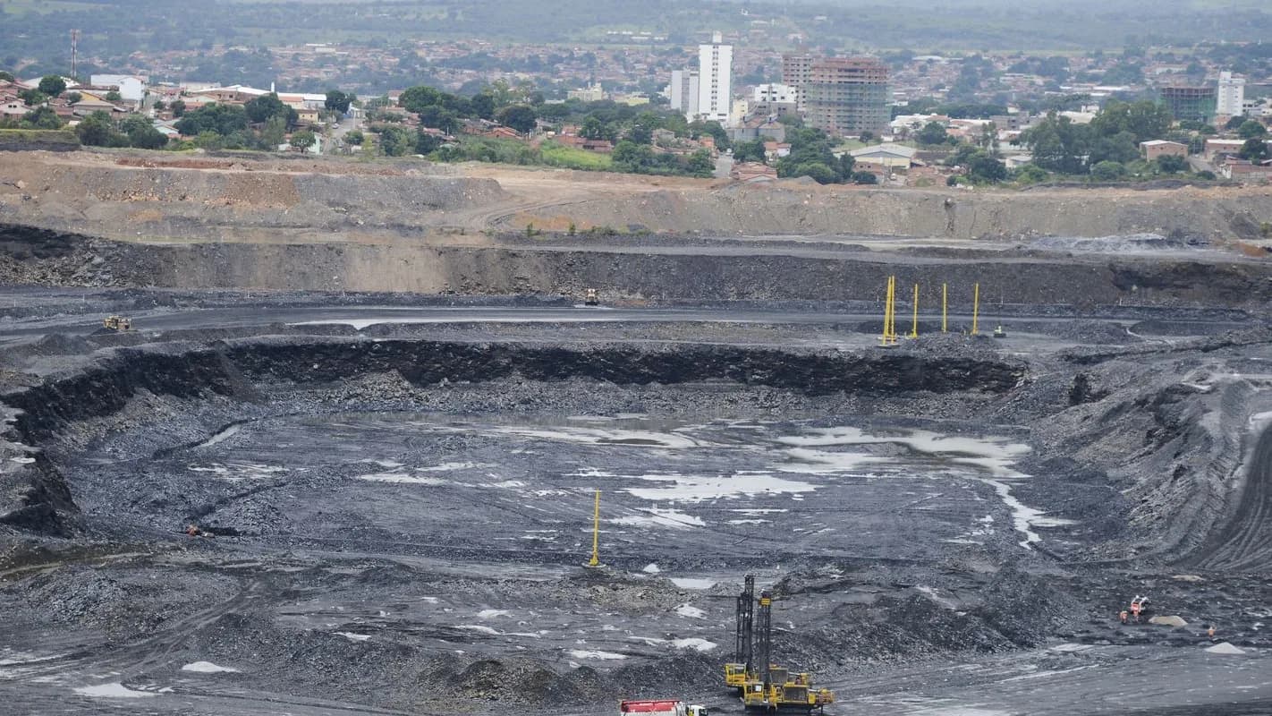 Vista aérea de mina a céu aberto no Brasil, com maquinário e caminhões de mineração ao fundo de área urbana