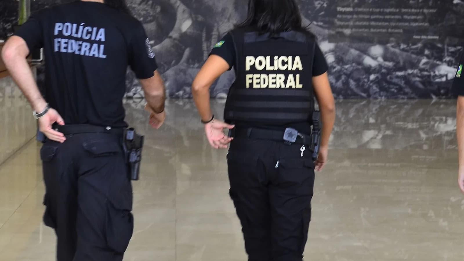 Agentes da Polícia Federal, vistos de costas, com o colete e a camisa identificados com o nome da corporação