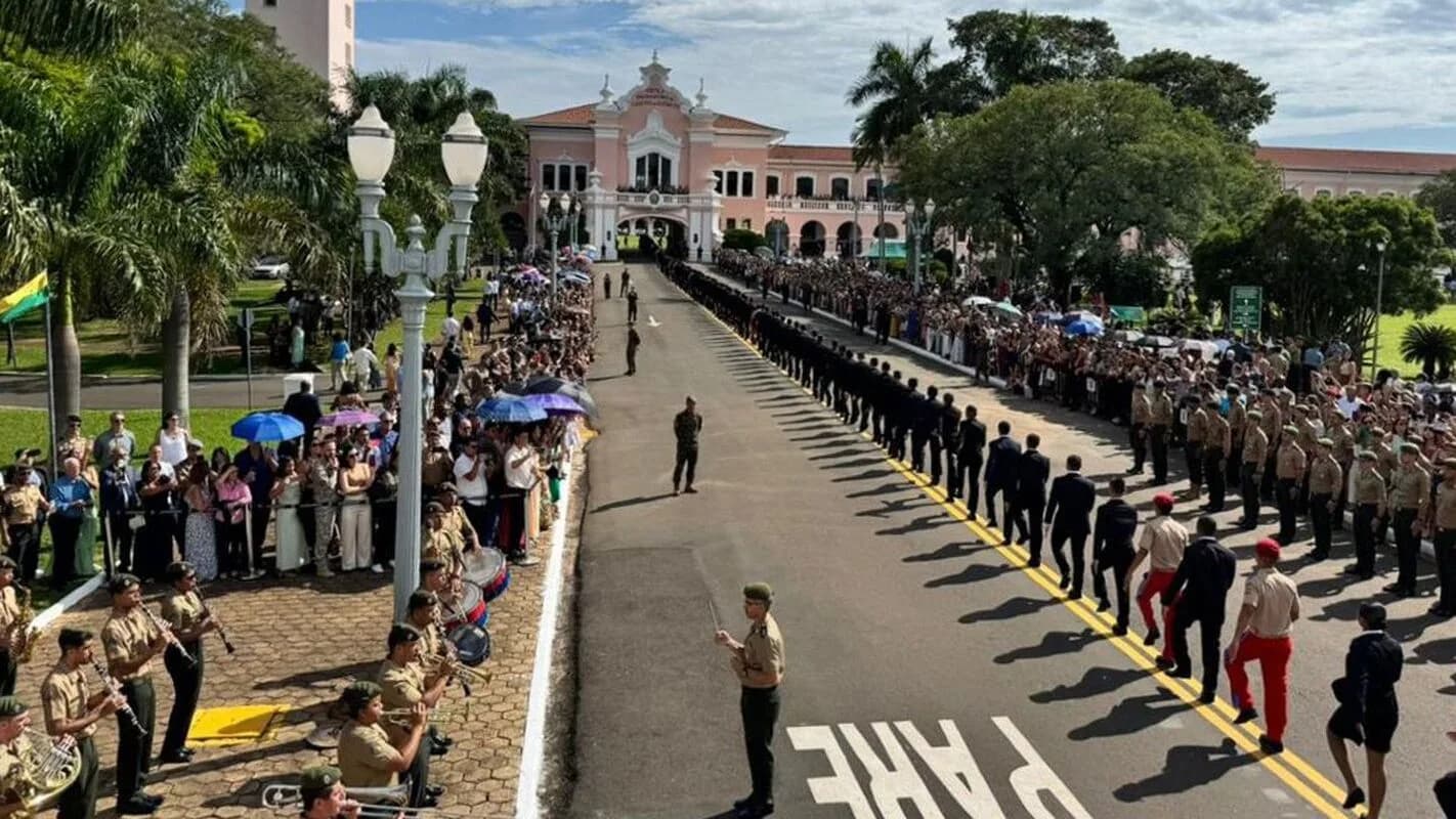 Cadetes e banda marcial da EsPCEx desfilam em avenida arborizada diante do prédio histórico da escola, com público observando dos dois lados