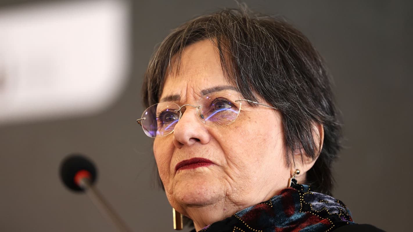 Maria da Penha, de óculos e cachecol estampado, em evento público falando ao microfone