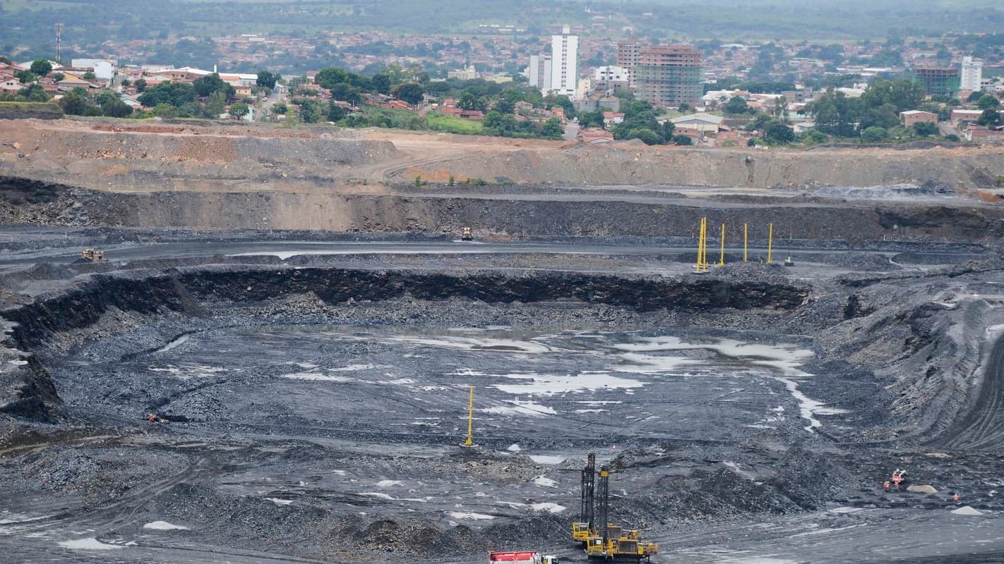 Vista de ampla mina a céu aberto com bancadas escavadas, sonda de perfuração e máquinas ao fundo de uma área urbana; imagem de arquivo