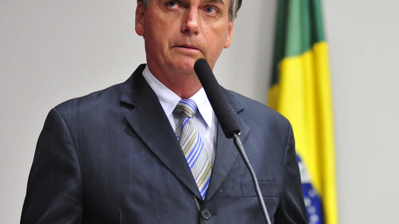 Retrato de Jair Bolsonaro em pronunciamento oficial, com bandeira do Brasil ao fundo