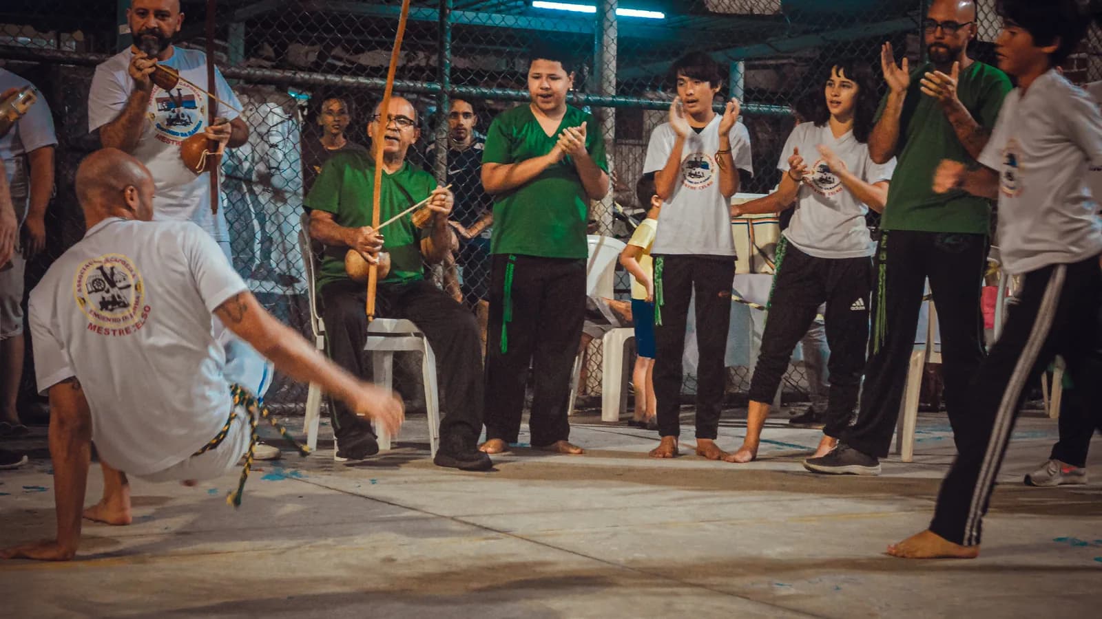 Roda de capoeira com jogadores em movimento e público em volta, com berimbau ao fundo