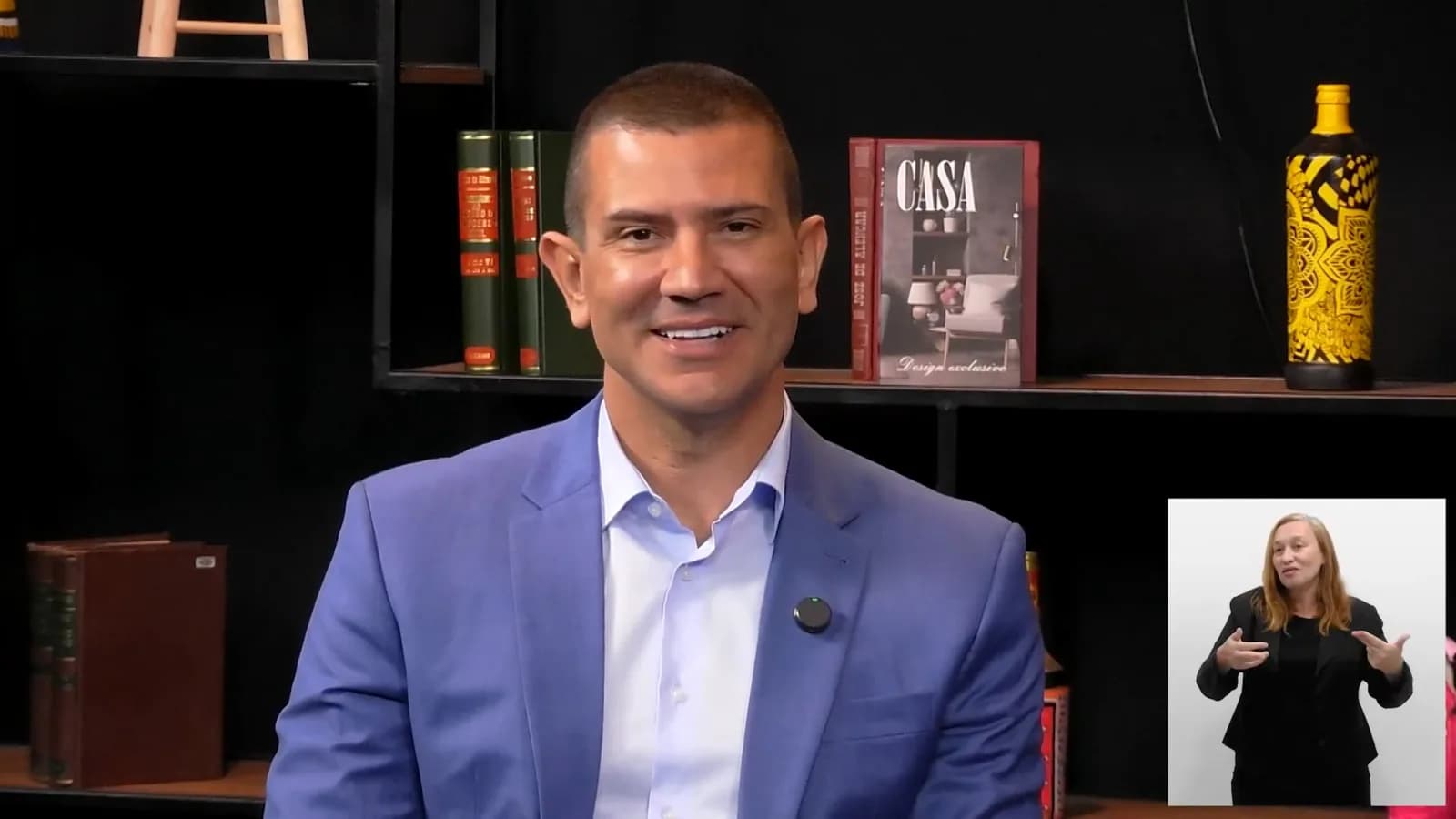 Douglas Ruas sorri durante entrevista em estúdio de TV, vestindo blazer azul