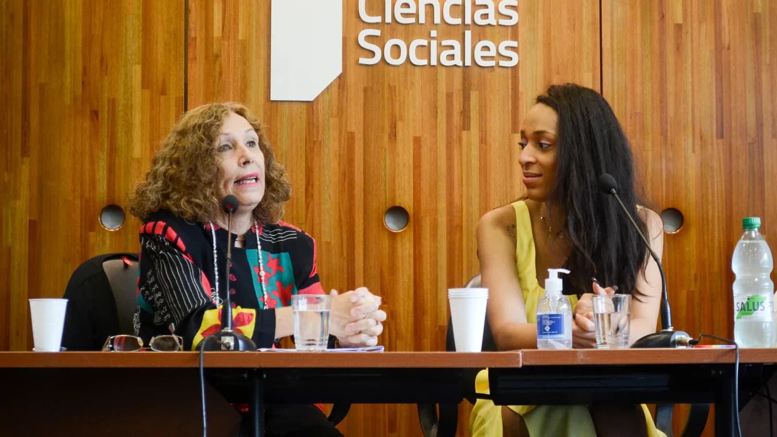Érika Hilton em conversatório público, sentada à mesa ao lado de outra palestrante