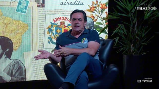 Domingos Sávio gesticula com a mão próxima ao peito, usando pin da campanha na camisa polo azul-marinho, durante a entrevista.