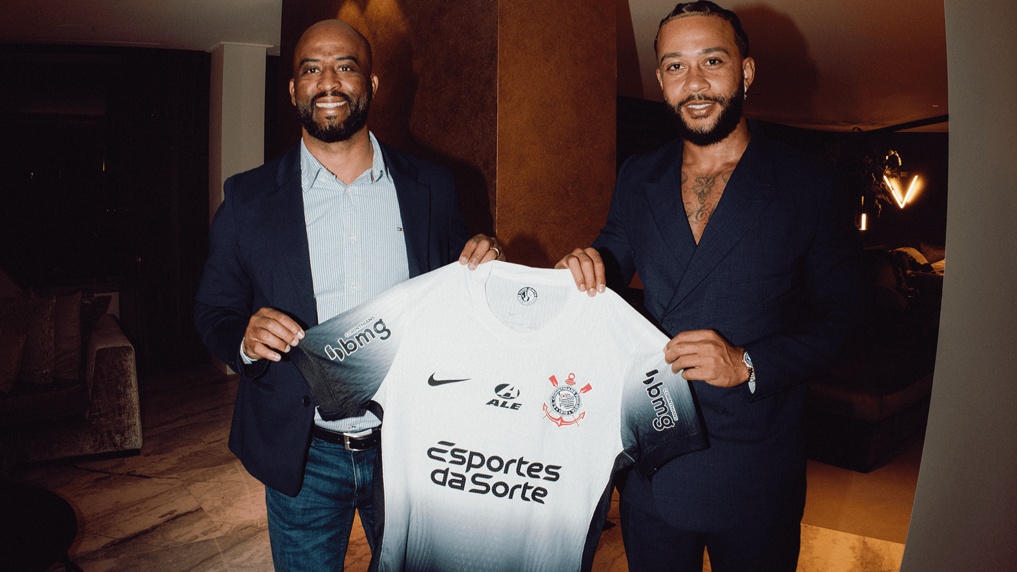 Memphis Depay ao lado de dirigente do Corinthians, segurando a camisa do clube, durante anúncio de contratação em 2024