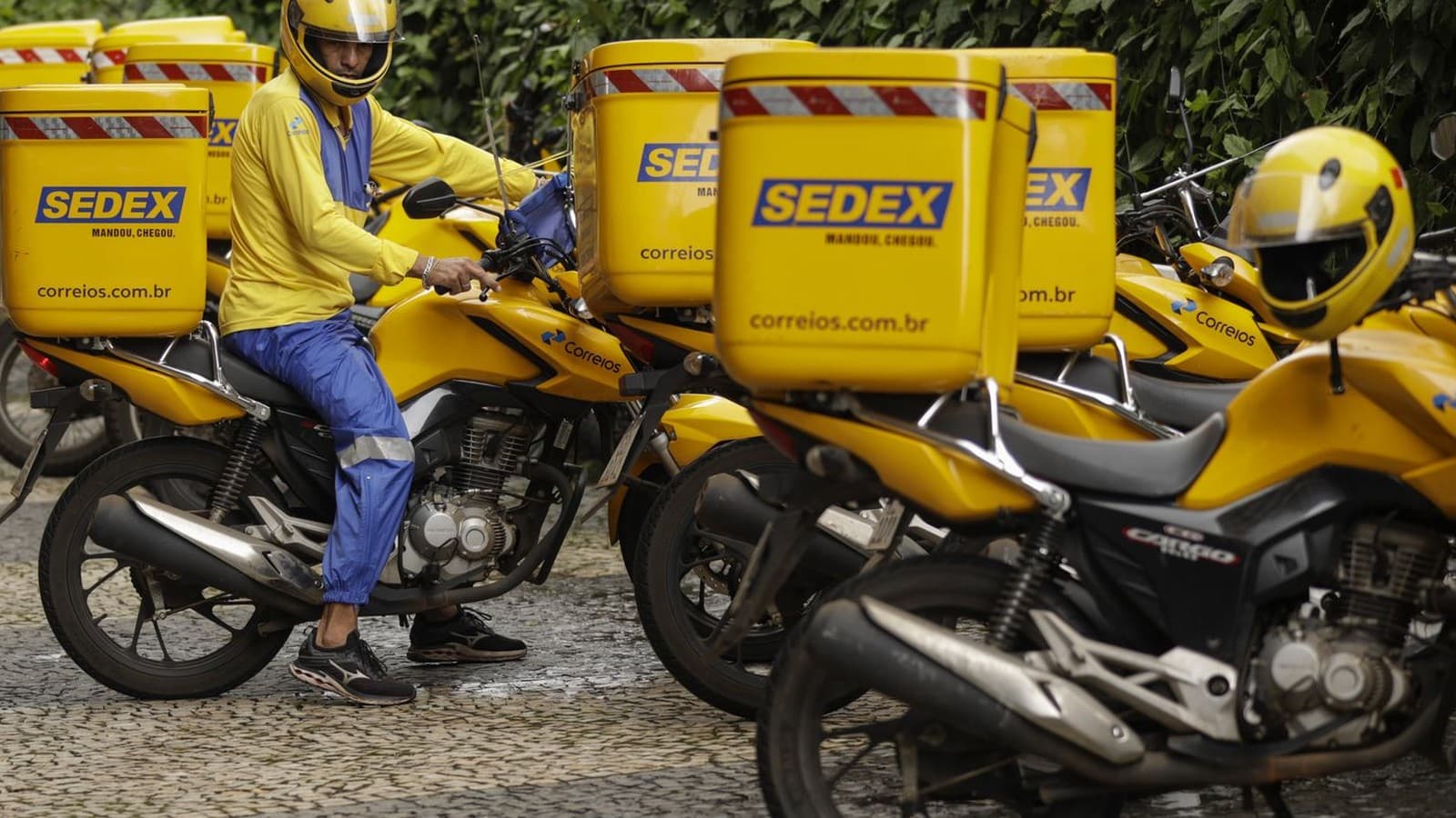 Carteiro dos Correios, de capacete e uniforme amarelo, sobre uma moto cercada por baús do Sedex, em imagem de arquivo